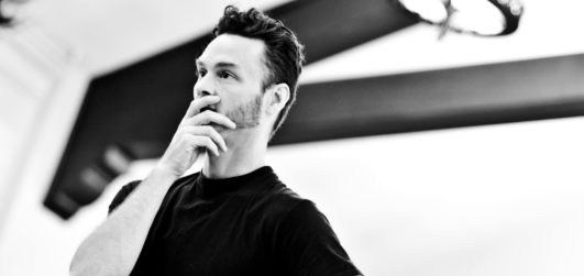 A black and white image of a male presenting person, Jonathan, stands with his hand on his chin looking on at a rehearsal.